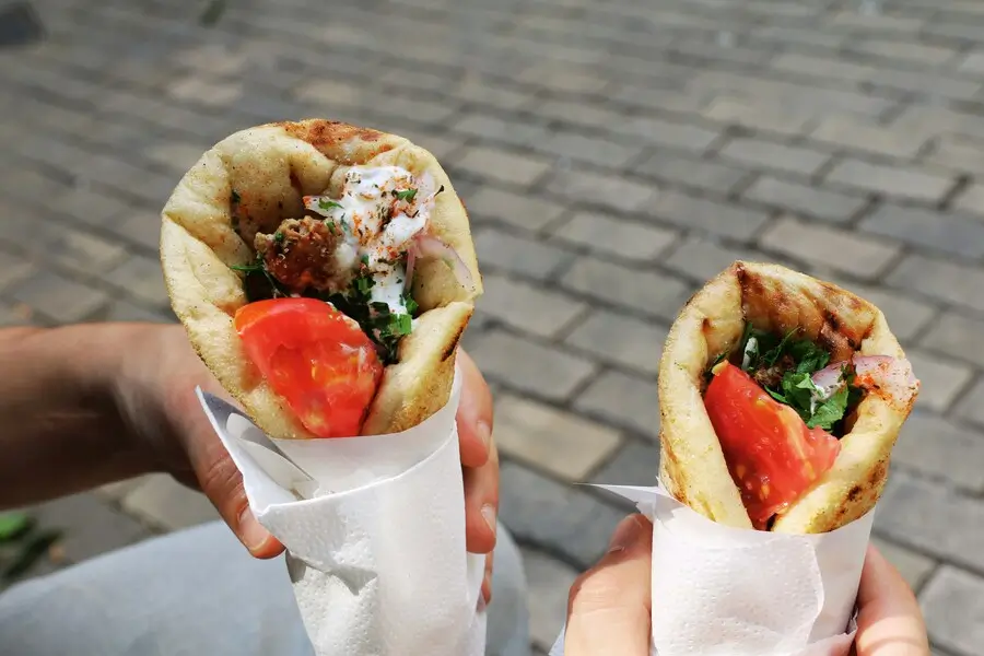 Two hands hold pita wraps with falafel, tomatoes, and lettuce on a cobblestone street.