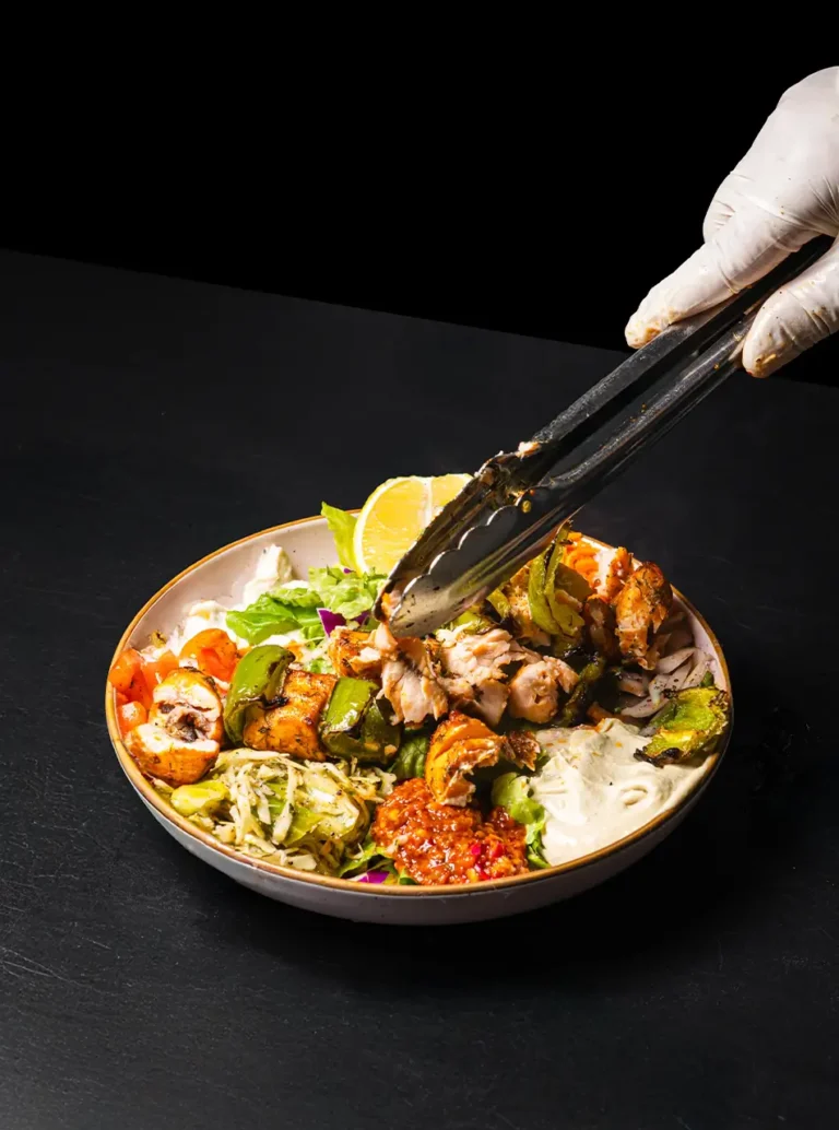 Chicken shish kebab bowl with green peppers, hummus, salad and carrots being served with tongs
