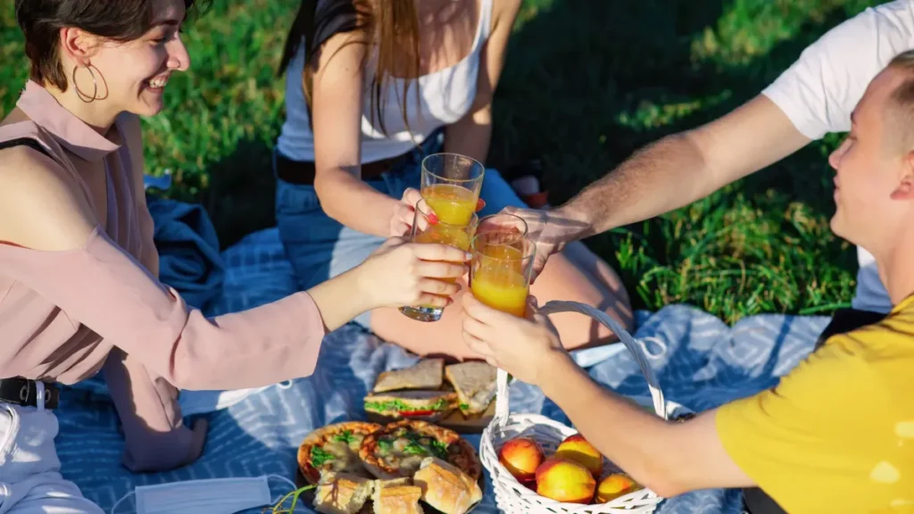 Friends toasting orange juice at an outdoor picnic with sandwiches and fruit