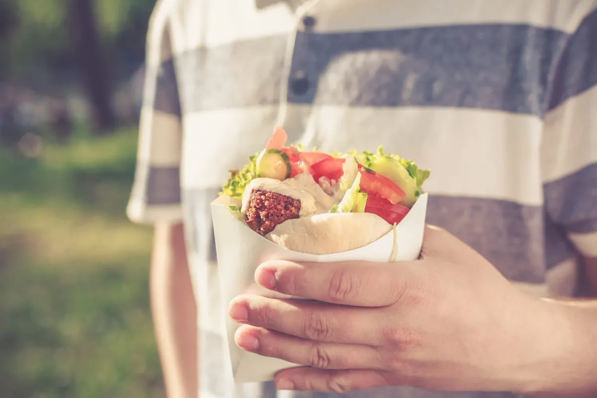 Hand holding a falafel pita wrap with tahini, tomato, cucumber, and lettuce outdoors