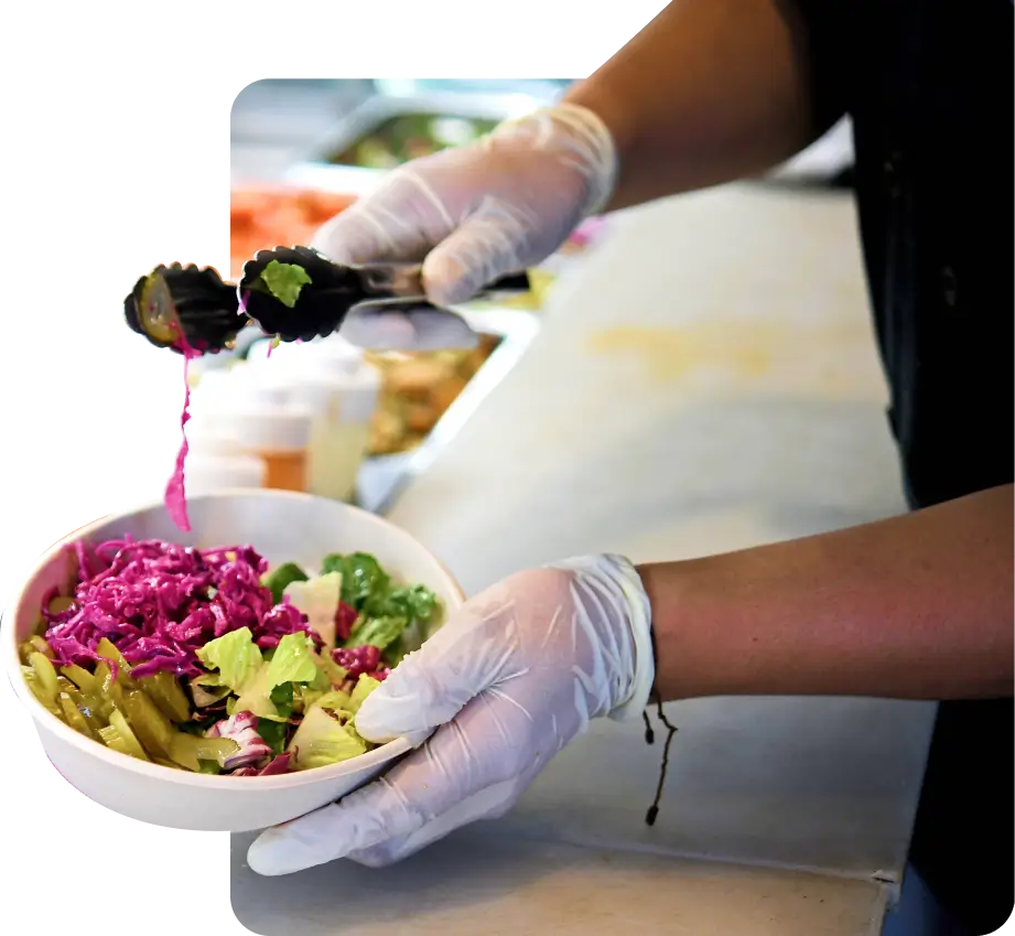 Gloved hands using tongs to add pickled red cabbage to a bowl of fresh greens and pickles