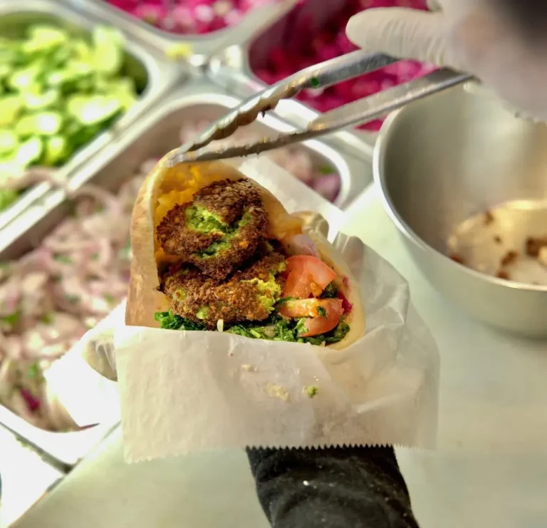 Falafel pita with parsley, tomato and greens being built at the topping bar with tongs