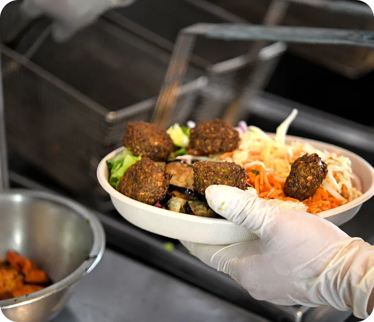 A gloved hand holds a bowl of falafel, vegetables, and shredded carrots.