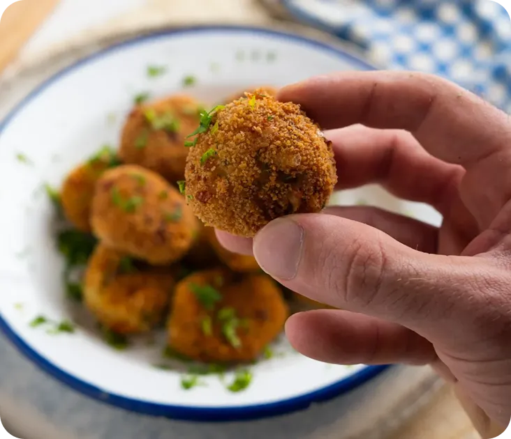 A hand holds a crispy, golden-brown fried ball over a plate of similar balls garnished with herbs.