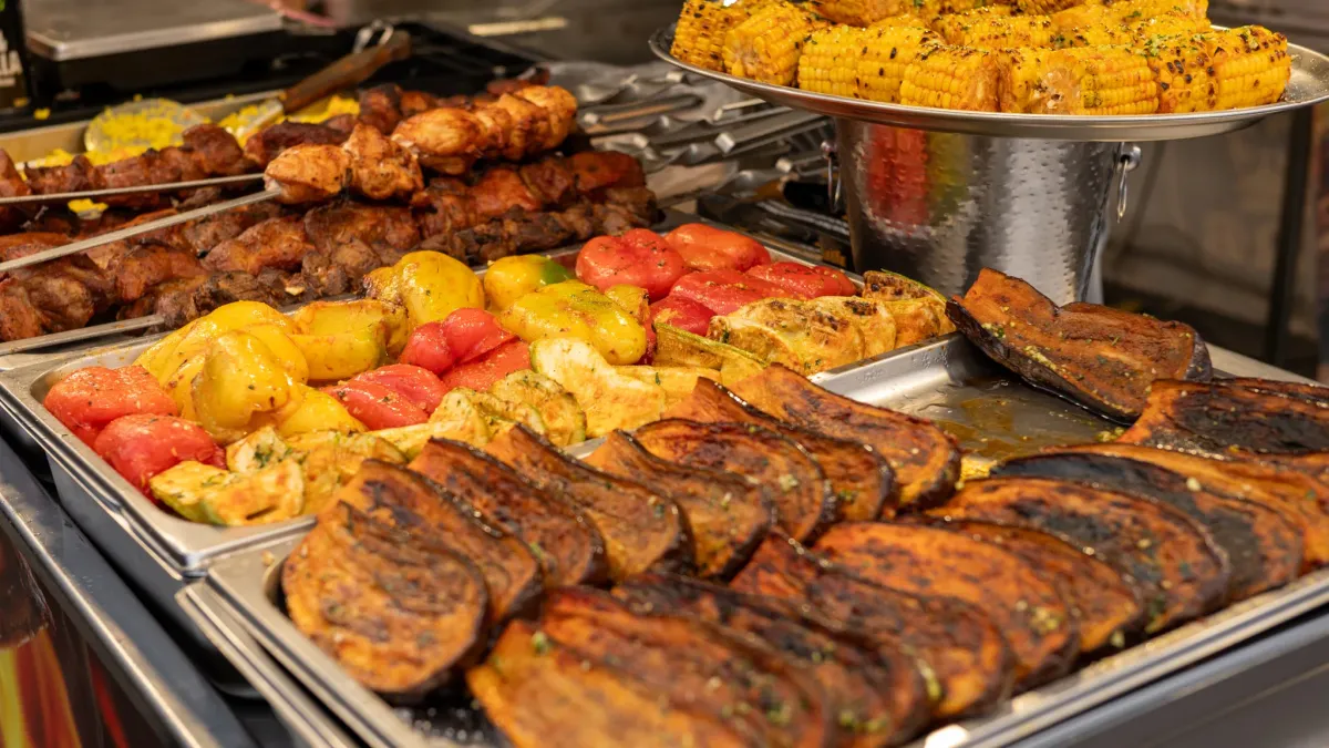 Buffet trays of grilled meat skewers, eggplant, peppers, potatoes, and corn