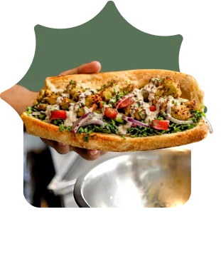 Hand holding a falafel sandwich with tahini, parsley, tomato, and red onion in flatbread